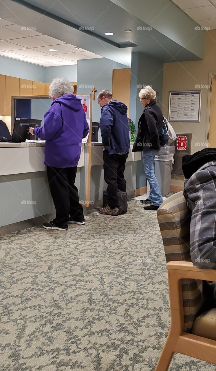 Men & women checking in at hospital reception desk. There is a line and people wait their turn quietly together. Others are already sitting while they wait to be called.