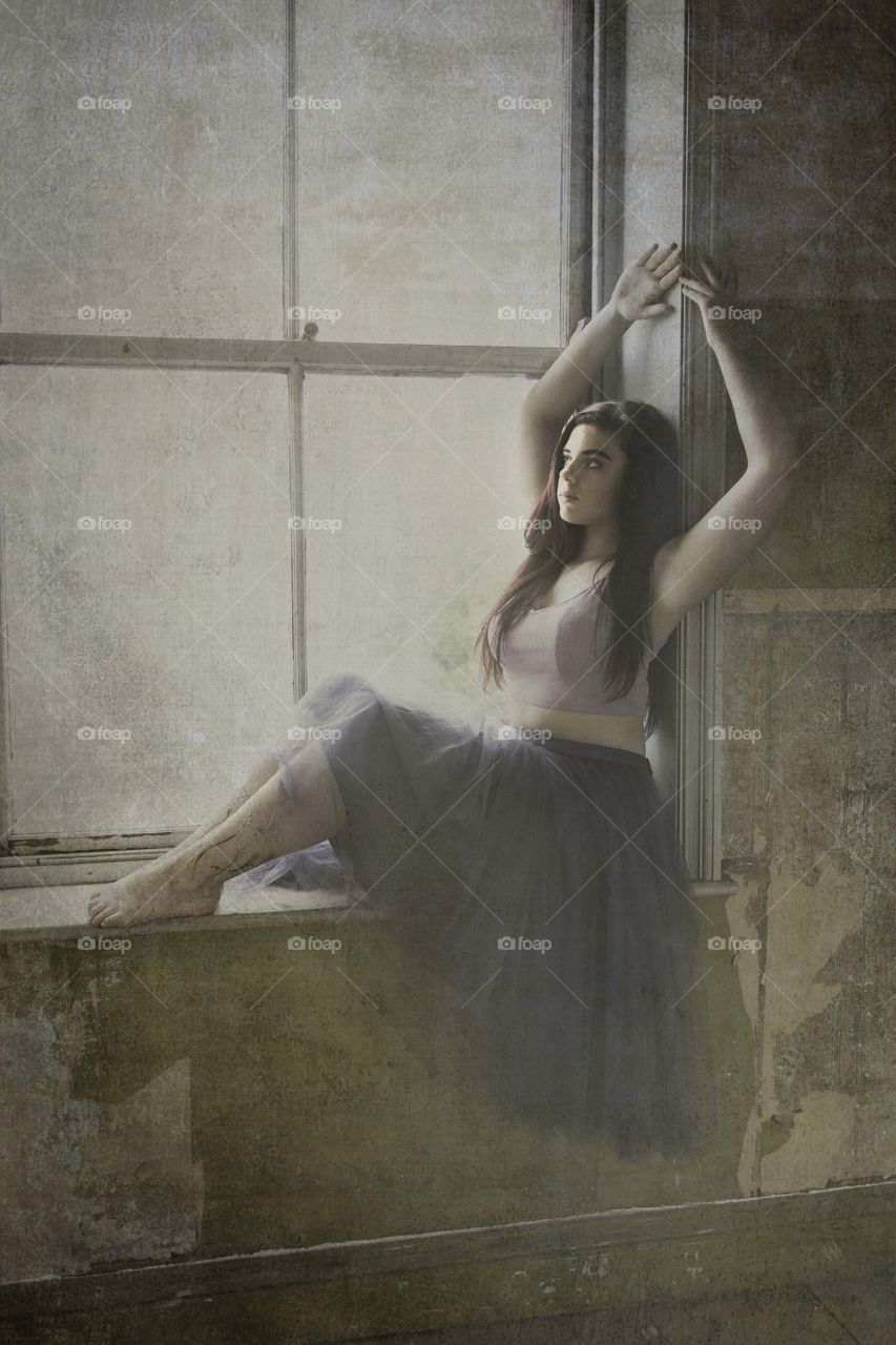 Girl seared on a window sill leaning against the wall she has her hands raised above her head. A texture has been added to the photo