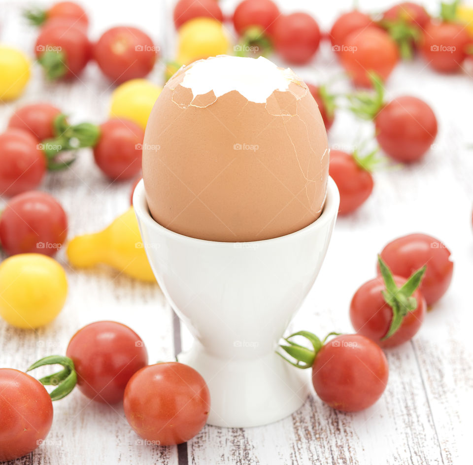 A hard boiled egg  in a white egg cup with red cherry ans tiny yellow tomatoes.