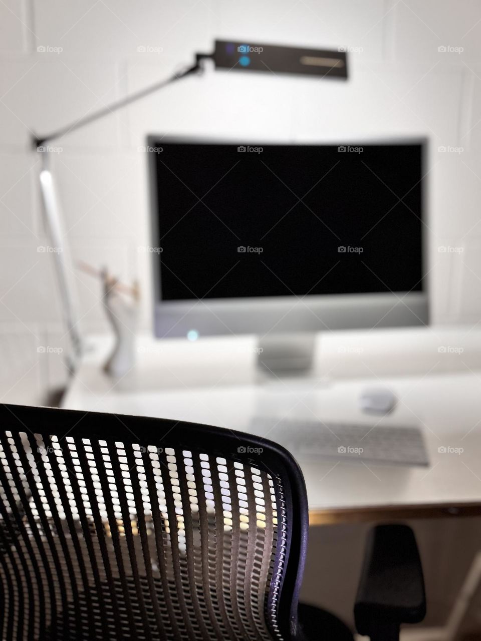 Drafting table desk with iMac, keyboard, mouse and lamp, and hand model with pencil, with black office chair in foreground 