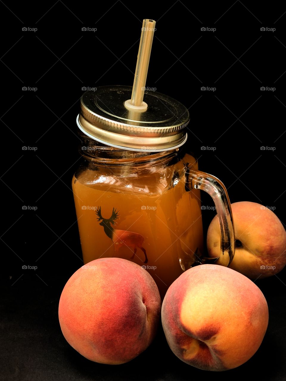 colorless mug with a handle and a metal lid with a hollow tube on a black background.  there is peach juice in the glass.  there are three peaches next to the glass