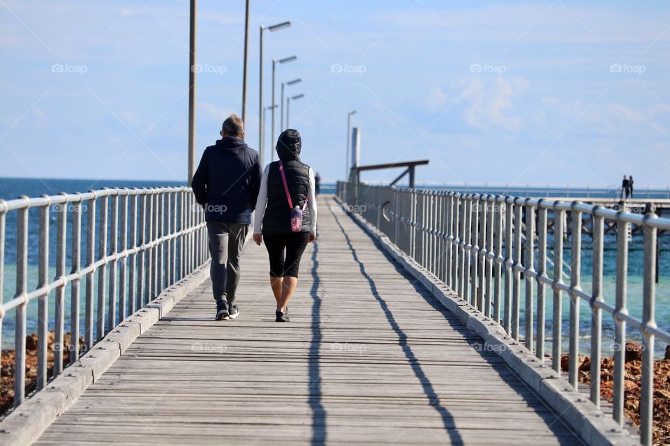 Man and woman couple walking along public ocean pier away from camera toward horizon 