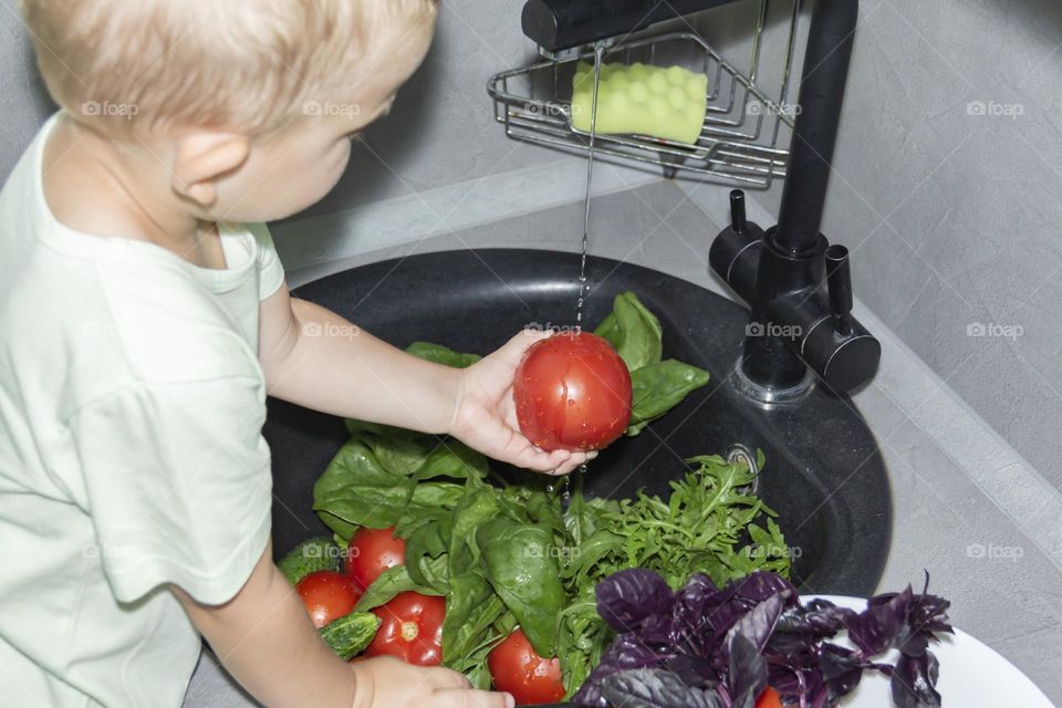 A small child washes fresh, green, vegetarian vegetables under the tap in a black sink to prepare salads and other dishes in a homemade gray and green kitchen.