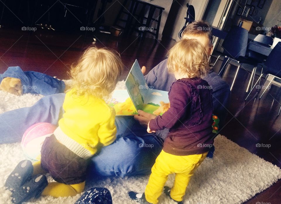 Coordinating identical twin toddler baby girls looking at book with uncle on floor
