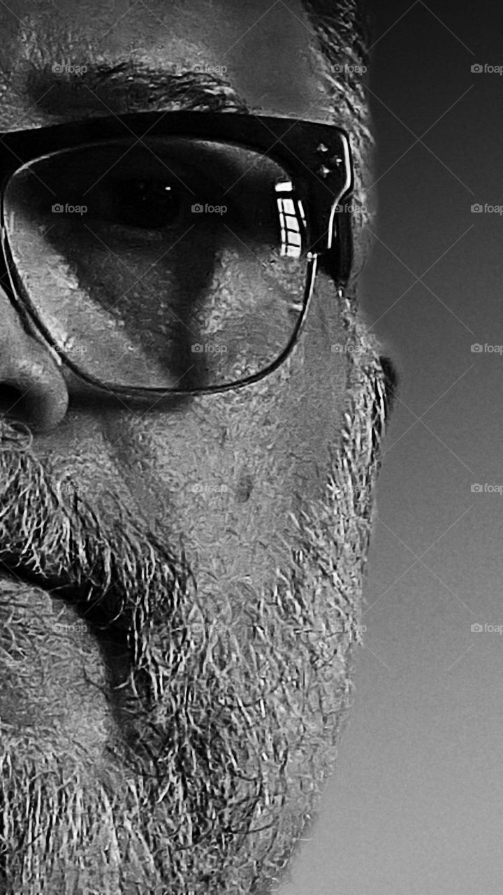 Half face of a man with a beard and glasses.  Reflection of part of the window in the eyeglass lens and shadows on the face.