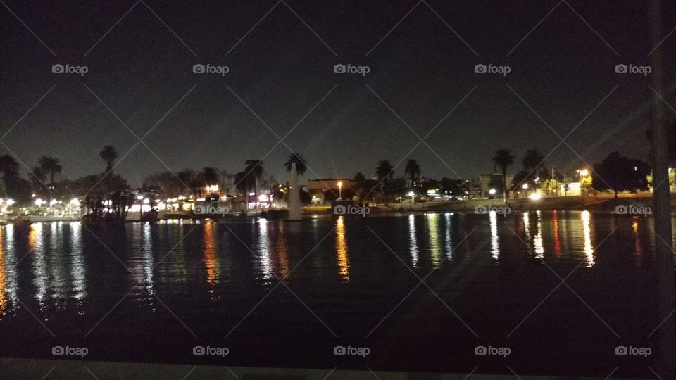 The reflection of the city lights on MacArthur Park Lake glow in rainbow colors. A fountain billows.