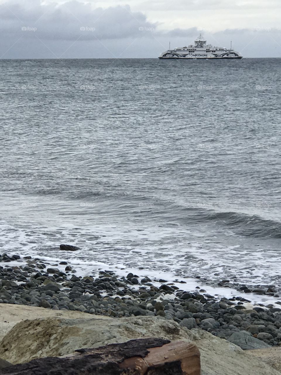A shot of the ferry crossing the Strait on a grey, blustery day. The stony, rocky, beach is in the foreground. The ferry is in the distance and behind it are grey and white clouds.
