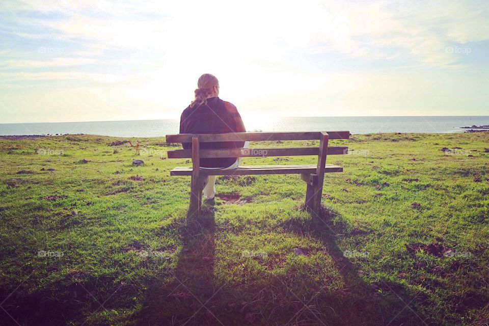 Man sitting alone on a wooden bench looking out at the clear blue sea on a sunny day at late summer