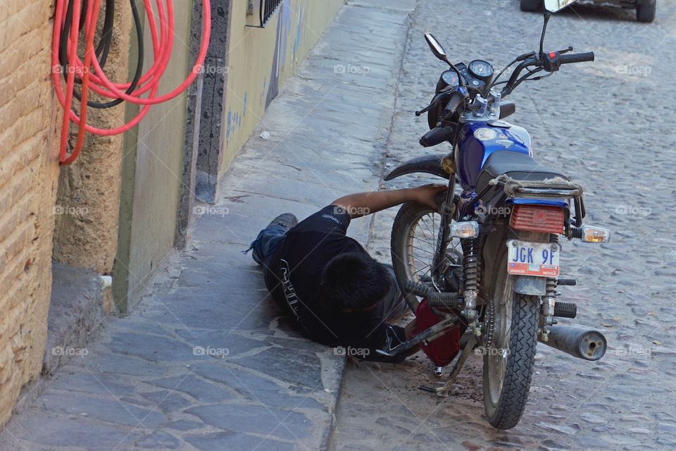 A young man, part of a father and son drive up auto and motorcycle street repair shop is repairing a motorcycle while lying under the vehicle on the sidewalk and in the gutter of San Miguel de Allende, Mexico,