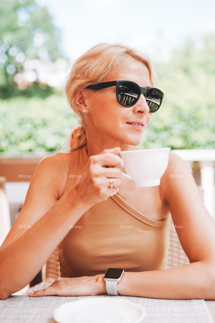 Adult elegant blonde woman in sunglasses beige clothes with cup of coffee in summer city street cafe