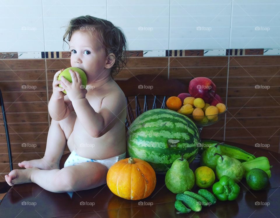 Still life. An adorable baby toddler holding a green apple. The baby encircled by summer fruits and seasonal vegetables. Crop. Natural light.