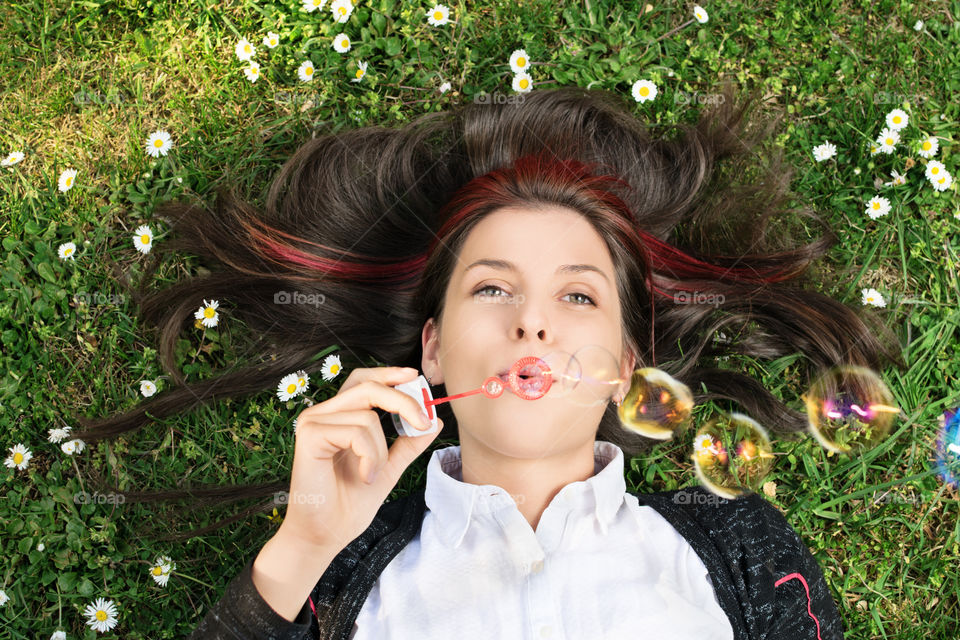 Lying on meadow's grass, making wishes and blowing soap bubbles in the air. Close up shot of a beautiful young student girl lying on a meadow blowing soap bubbles.