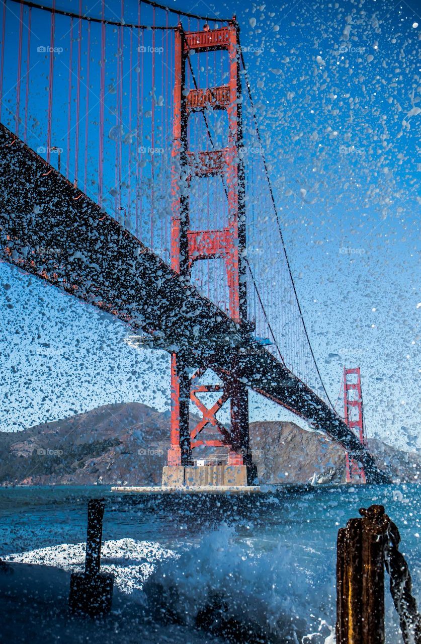 The Golden Gate Bridge seen through the splash of a wave as it hit at the base of fort point on a clear day in San Francisco California with the Marin Headlands in the background 