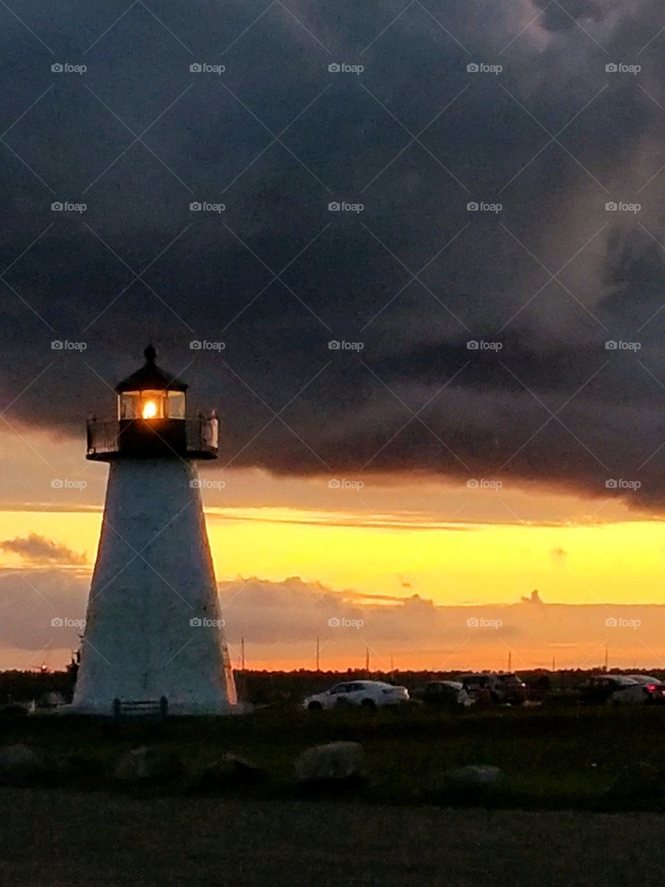 Lit lighthouse overlooking an oceans bay, guiding boats from shallow waters. Sunset/sunrise is colorful on the background horizon.