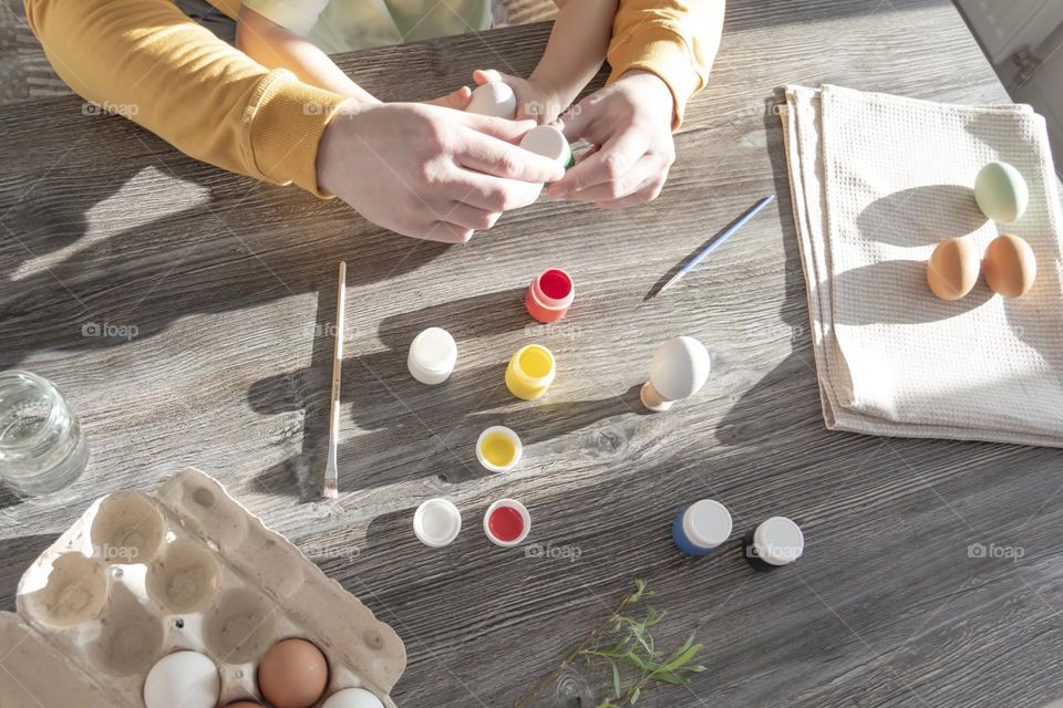 A little son and his father together in their free time paint Easter, chicken eggs with paints and brushes for the holiday.  A child and a man create a craft and natural product in their family business.