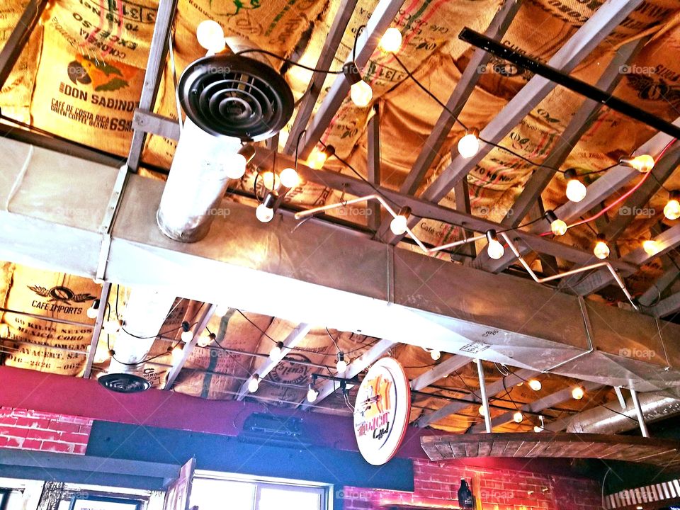 Urban warehouse interior designed space with exposed steel duct, beams, adorned with glowing string bulb lights for rustic ambiance with uniquely covered ceiling with floor sacks at neighborhood coffee shop.