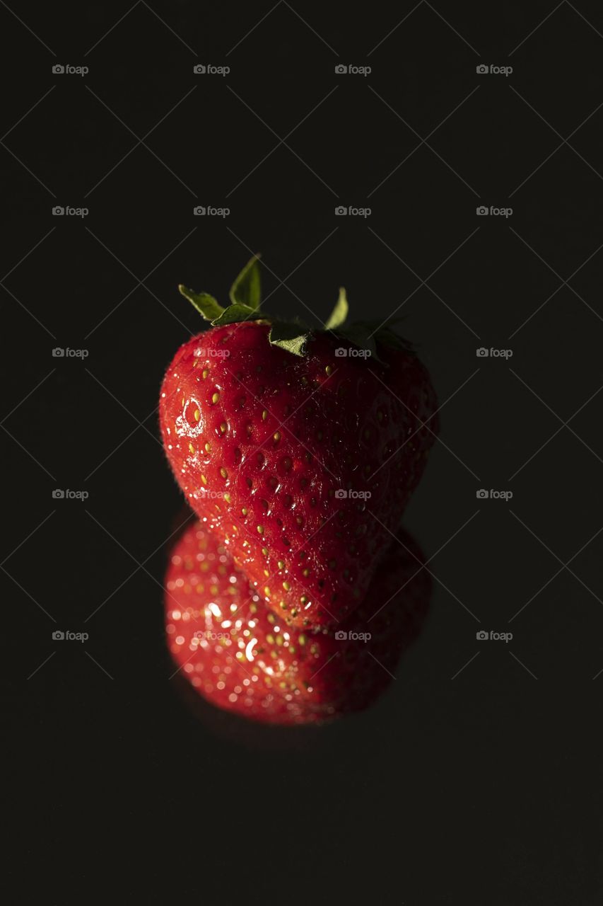 A portrait of a red fresh and healthy strawberry. the poece of fruit is being reflected by a mirror below it and is getting lit at the side.