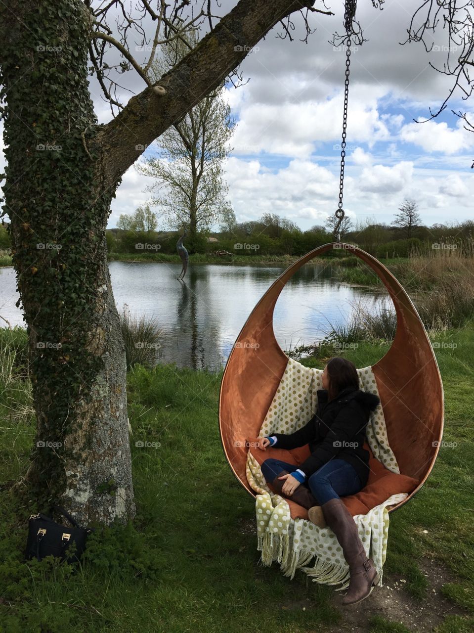 Girl on golden swing looking out to the beautiful scenic view of water, woodland and cloudy blue sky’s. Girl relaxing on swing chair by lake