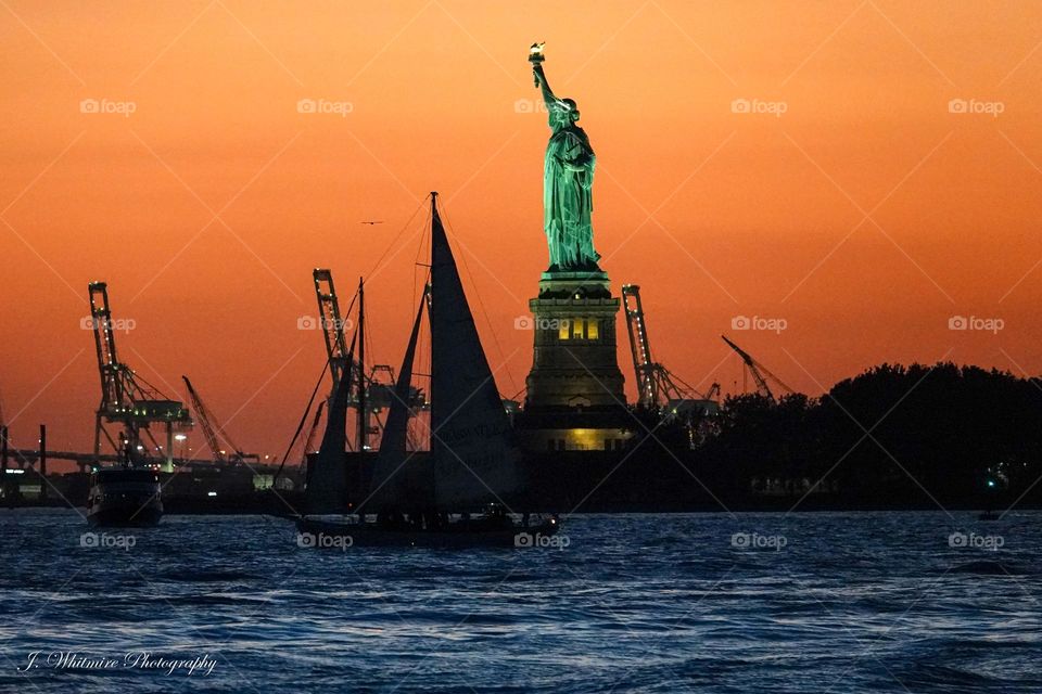The Statue of Liberty glows brilliantly in New York harbor in front of a vibrant sunset