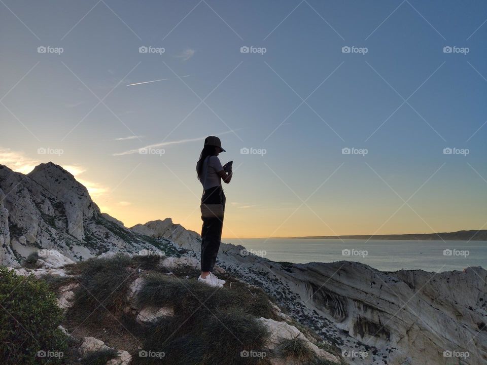 A portrait of one young Caucasian girl stands half-turned on a rocky cliff and takes a photo of the landscape on a smartphone at sunset on a summer evening, close-up side view.