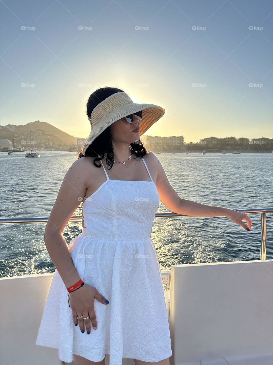 Woman standing on a boat, while wearing sunglasses and a hat, with the sunset as the background.