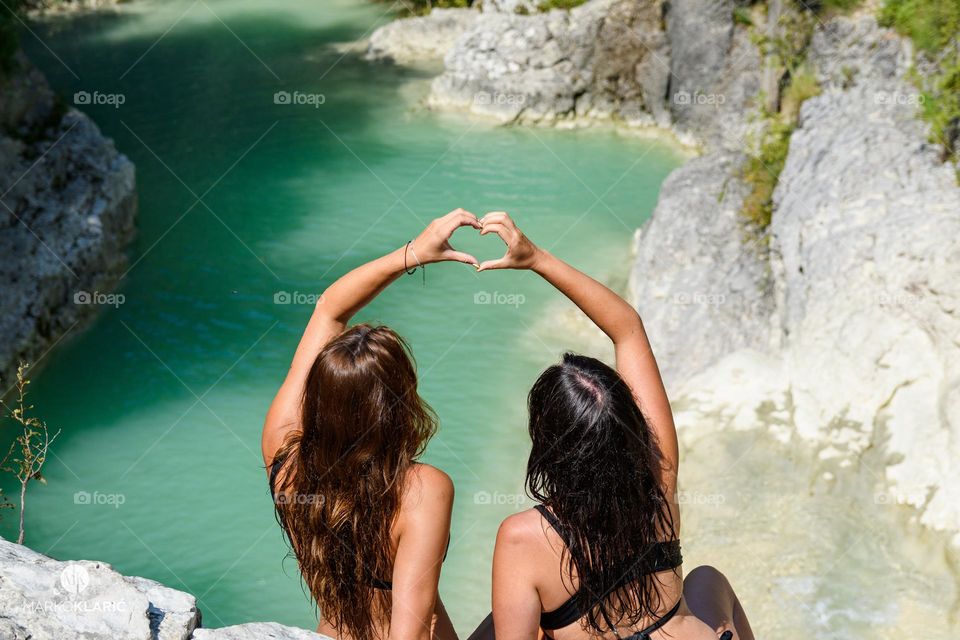 Back view of female friends in bikini sitting above lake, holding hands in shape of a heart