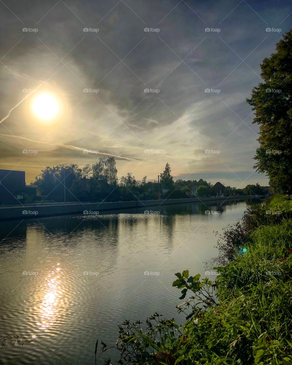 Sun setting or rising behind thick dark grey clouds over the reflecting water of a river or canal