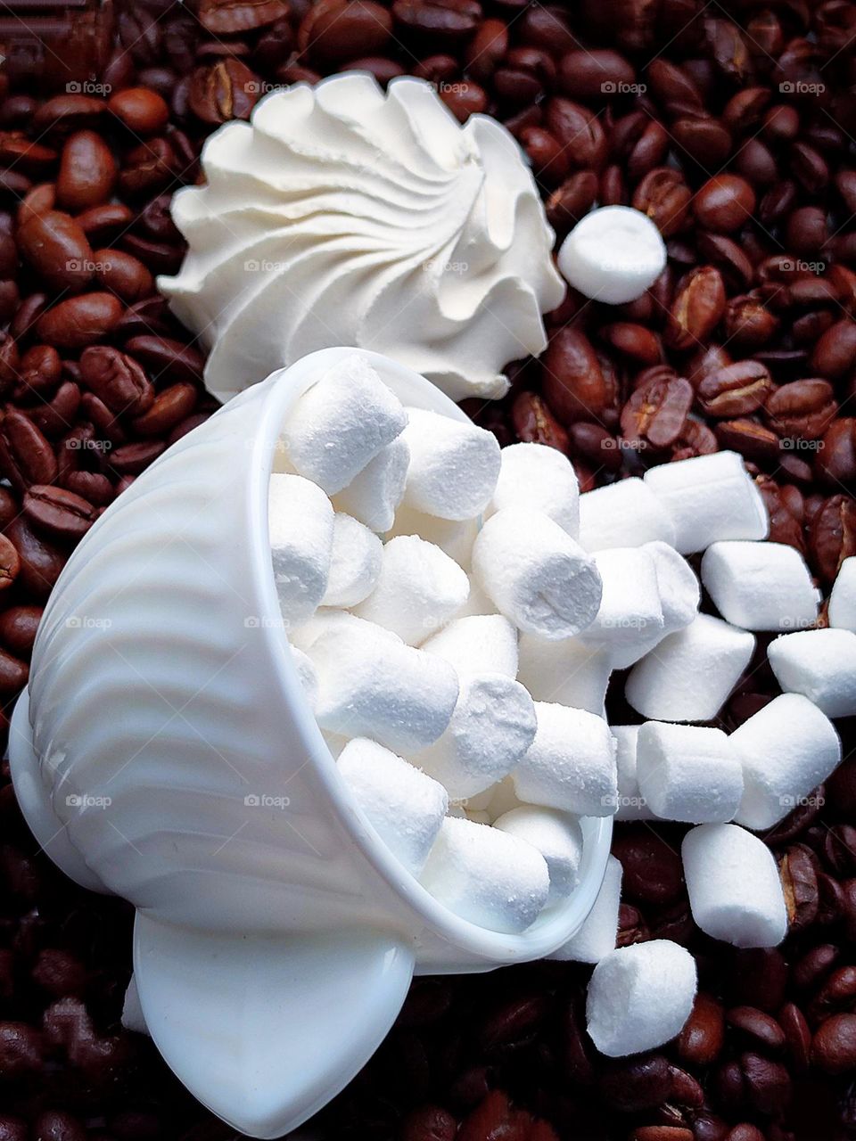 White colour. On brown coffee beans lies an overturned white cup with a white marshmallow that has spilled out of the cup. In the background lies a white marshmallow in the shape of a seashell. Contrast