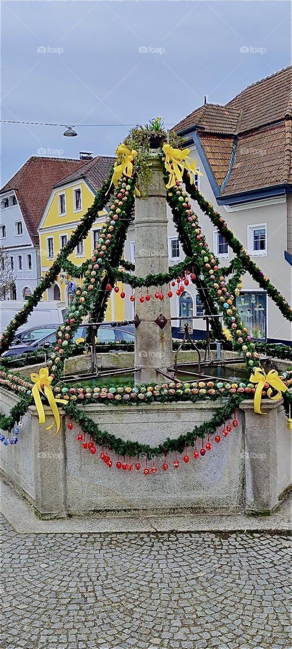 This old well on the town square of “Ortenburg” in “Bavaria”, Germany has been decorated traditionally for “Easter” with plant girlands, ribbons and artificial painted eggs. 2024. Hypnotic Productions