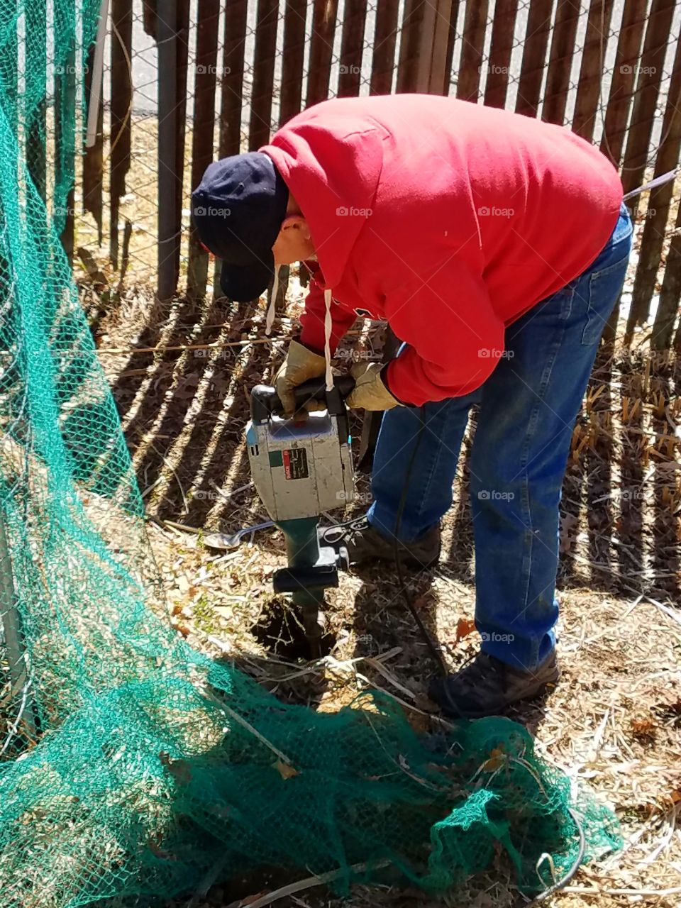 Worker jackhammer holes for future pipes, extending chain link metal fencing. Tool is in use in photo, noisy.
