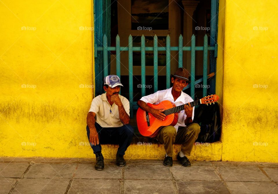 Busker and a man in the streets of Trinidad, Cuba on Christmas Eve 2013.
. A street musician and another man sitting by the wall of an old yellow building in the historic part of Trinidad, Cuba on Christmas Eve 2013.