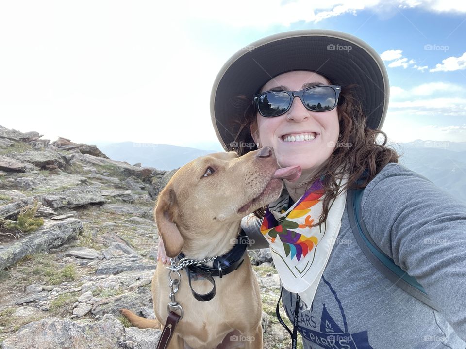 Dog licking a woman smiling on a mountain in nature, with sun hat, bandana and sunglasses. 