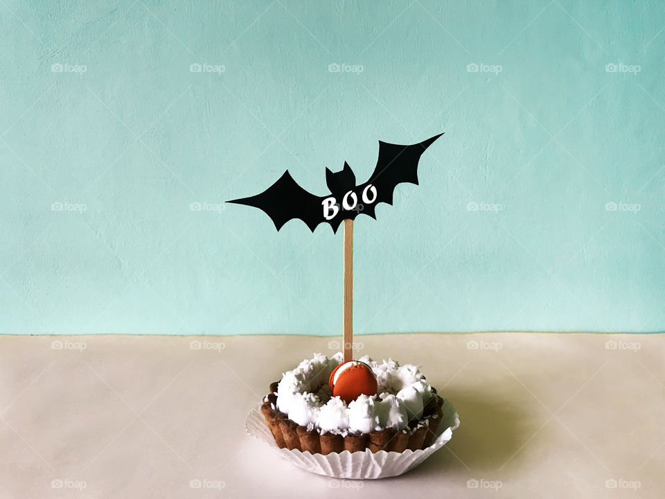 Festive cupcake with pumpkin macaroon and Halloween decor with a word “Boo” on monochrome pastel background 