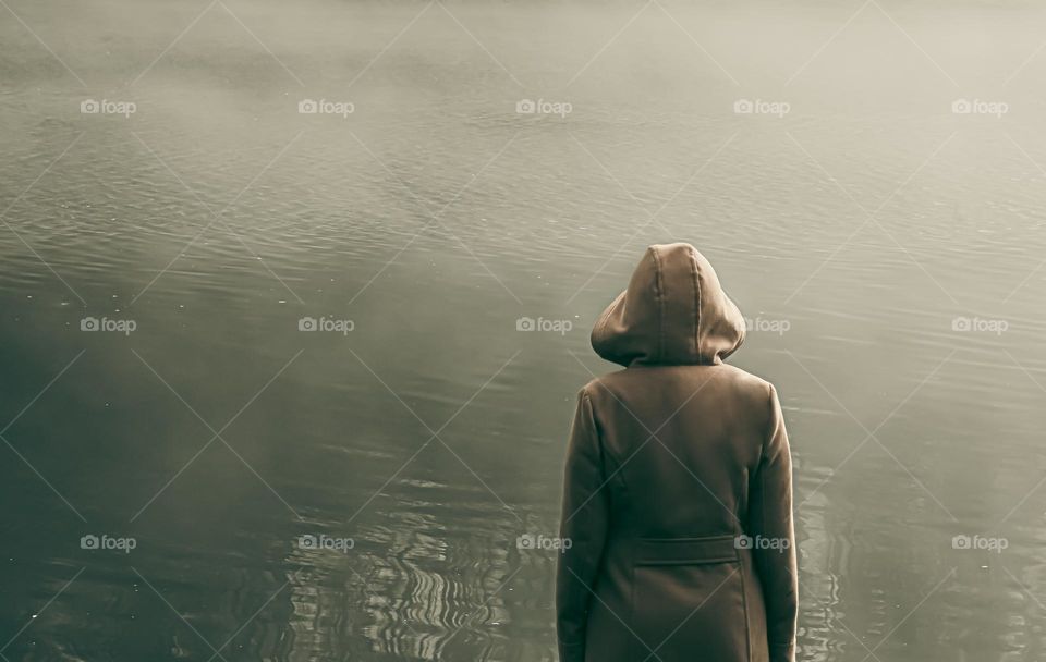 A hooded figure looks out over misty water.