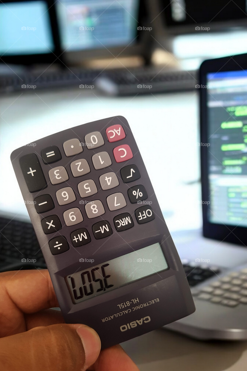A person holding a Casio calculator showing the word 'LOSE' on its display, with a computer and coding screens in the background. The image represents a mix of humor and technology in a work environment.