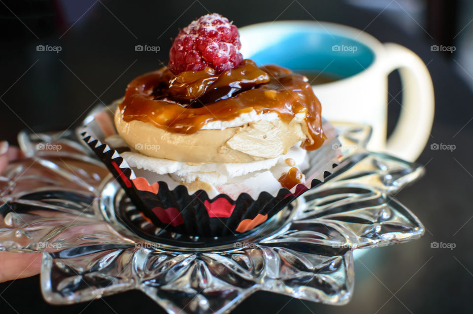 Holding a decadent caramel buttercream nest cupcake on crystal plate topped with a raspberry next to a cup of coffee on the table 