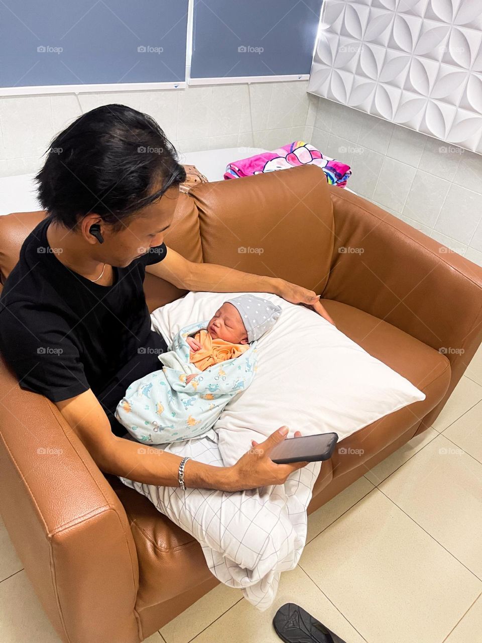 A father sitting on a brown sofa, holding a baby wrapped in a light blue blanket