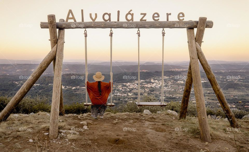A woman in a sun hat a orange shawl sits alone on the twin swings, around sun down, overlooking the town of Alvaiazere in Central Portugal 