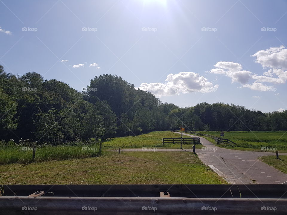 Uitzicht vanuit een auto, naast de auto weg ligt een fietspad. Met landschap er omheen, bomen en gras. De zon schijnt heerlijk in de lucht met een beetje wolken.