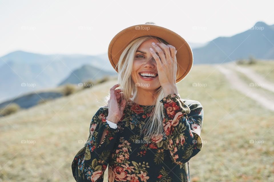 Beautiful smiling blone young woman traveler in dress and felt hat on road, trip to mountains