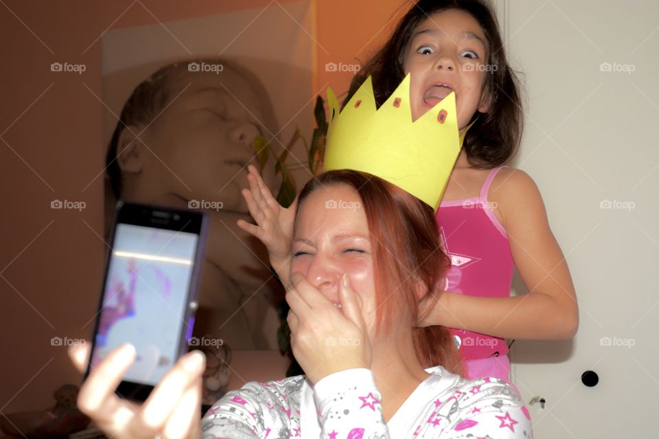Little girl fooling around with an adult woman, combing her hair and putting on a paper crown, adult woman laughs and takes a selfie but bursts into laughing