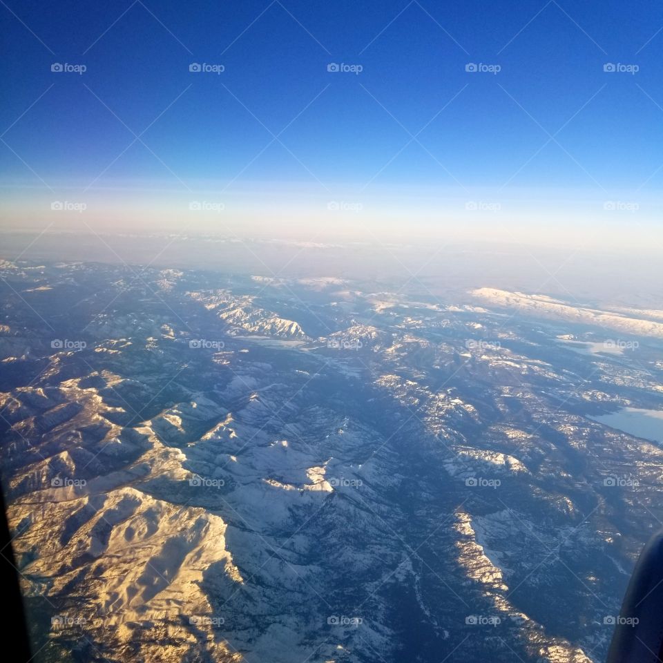 airplane view of snow covered mountain peaks with a glow of sunshine  and blue sky