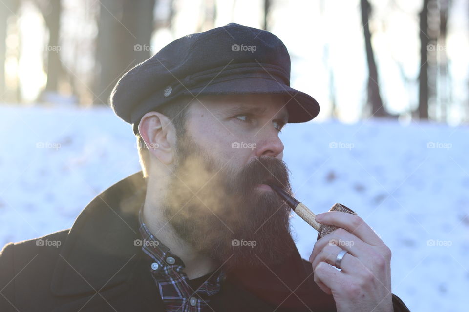 A middle aged bearded man smoking out of a corn cob pipe during winter.