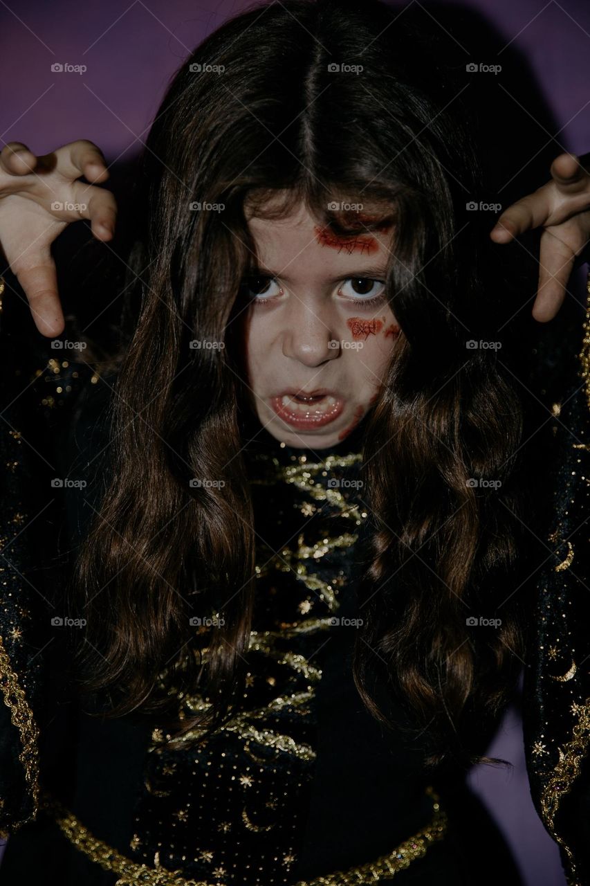 Portrait of one beautiful little Caucasian girl with bloody scars on her face in a witch dress, frightening with raised hands and terrifying emotions on her face, close-up side view.