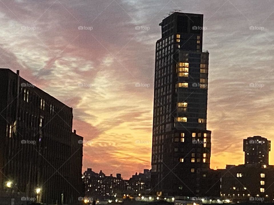 An imposing dark silhouette of a high rise building towers over the waters of the East River at Newtown Creek, Queens, NY just as the sun sinks below the horizon. 2021. Hypnotic Productions