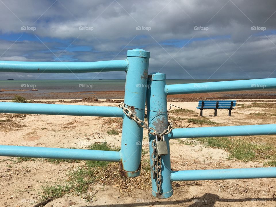 Aqua marine blue locked metal gate with chain and padlock barring entrance to beach and ocean on cloudy day 