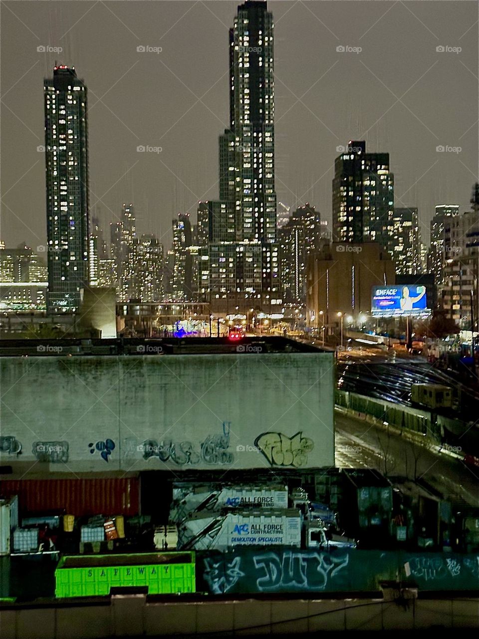 This is a view from the staircase of the “Pulaski Bridge” at “Newtown Creek” in LIC, Queens overlooking adjacent buildings onto the tracks of the “Long Island Railroad”, the LIC skyline and further back “Manhattan”. 2023. Hypnotic Productions