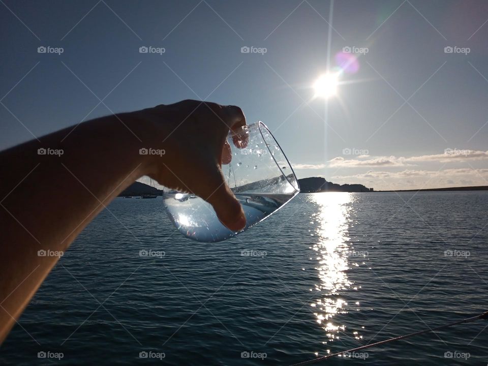 Juxtaposition of a Glass of Water being held and tipped and lined up with the Ocean Horizon on a lovely Sun lit evening in Sassari Province, Sardinia. The Sea is Grey Blue and shimmering like Stars and Diamonds. The sinking Sun reflects on the Sea.
