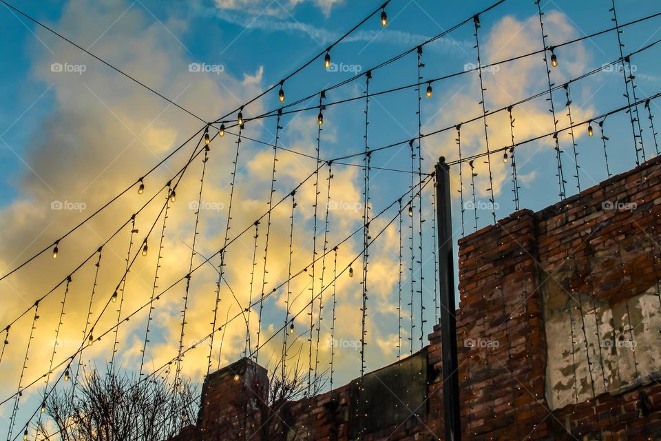 View of a bridge from below . Clouds in a blue sky , wires meeting with lights abound 