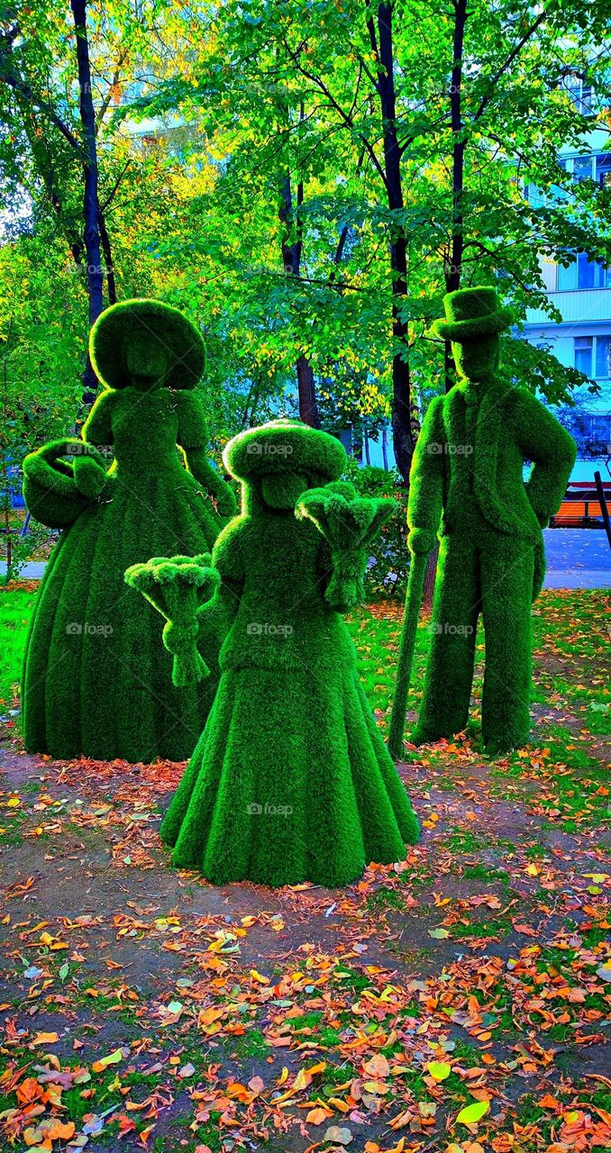 Autumn.  Three figures made of green artificial material stand on the asphalt strewn with colorful fallen leaves.